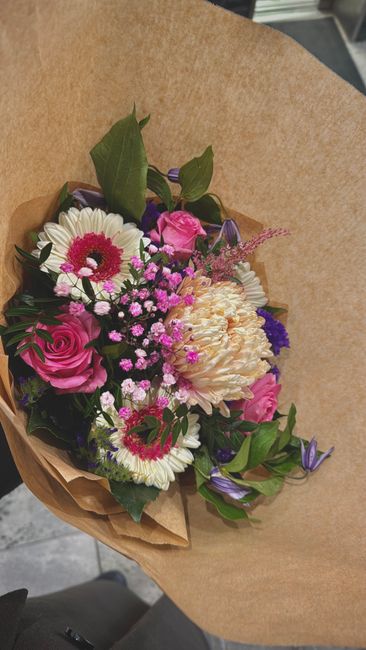 A bouquet of fresh flowers featuring pink roses, white daisies with pink centers, purple and pink waxflowers, and a large white peony, all arranged with green foliage. The bouquet is wrapped in simple