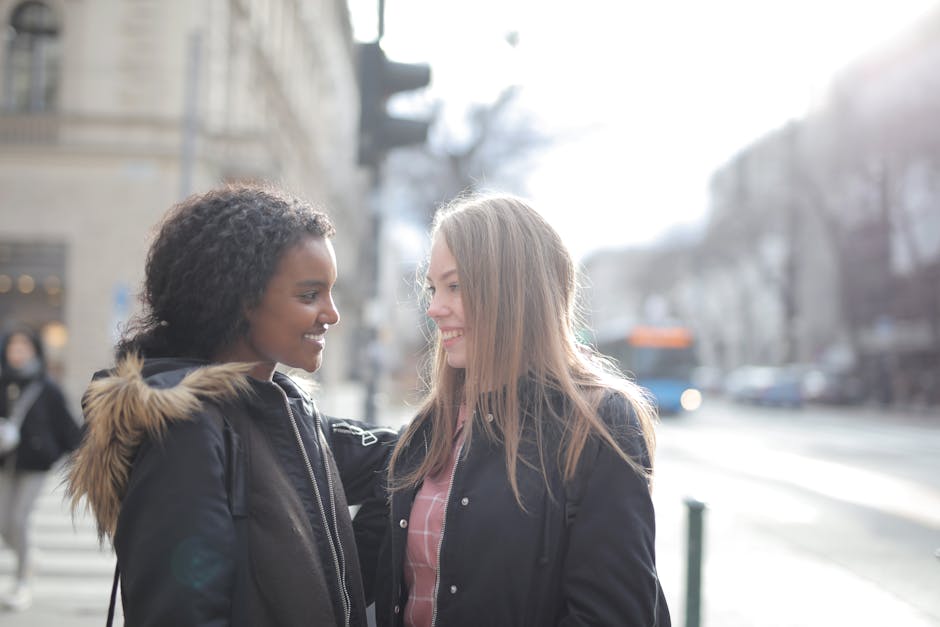 Two women smiling and facing each other on a city street, with the woman on the right holding a floral gift bouquet arranged in a clear glass vase. The bouquet features a variety of fresh flowers, inc