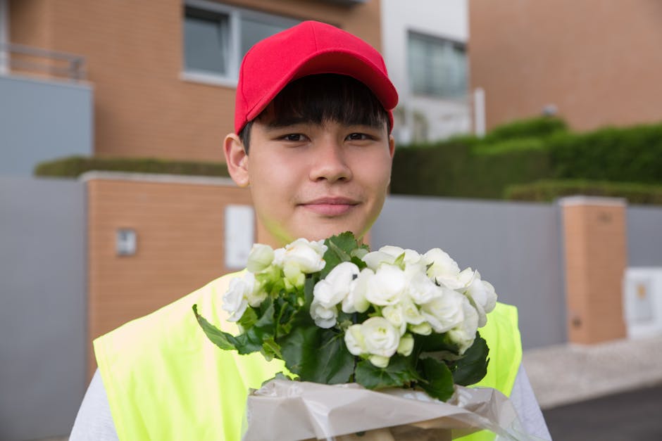 A young male flower delivery courier wearing a bright yellow high visibility vest and a red cap, holding a bouquet of white roses wrapped in white paper. He is standing outdoors in front of modern res