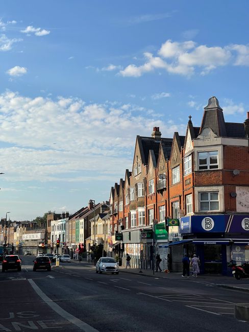 The image shows a street scene with a row of multi-story brick and stucco buildings featuring gabled roofs and bay windows, typical of urban British architecture. Several shopfronts line the sidewalk,