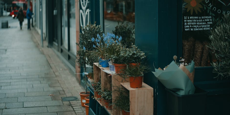 A display of potted flowering plants arranged on a wooden shelf outside a flower shop, with blue, white, and terracotta pots holding various blooms and greenery. The flowers include small blue flowers